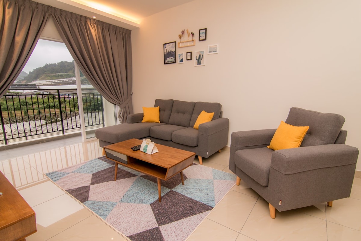 A living room space features a comfortable gray sectional sofa and two matching chairs, accented with yellow cushions. A coffee table sits centrally on a multi-colored rug. Large windows with gray curtains provide a view of the surrounding landscape.