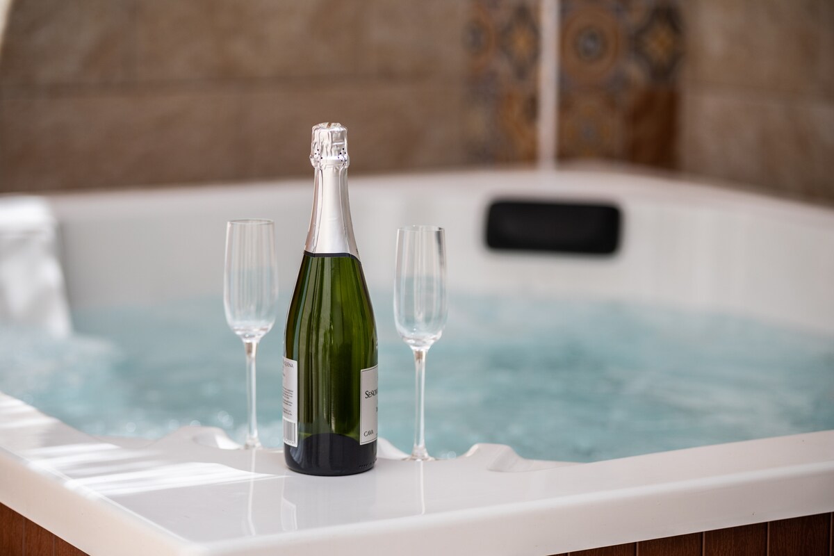 A spa-like setting is highlighted by an inviting Jacuzzi filled with bubbling water. A bottle of champagne sits upright beside two elegant flutes, creating a perfect atmosphere for relaxation. The surrounding tiles feature a subtle decorative pattern, enhancing the tranquil ambiance.