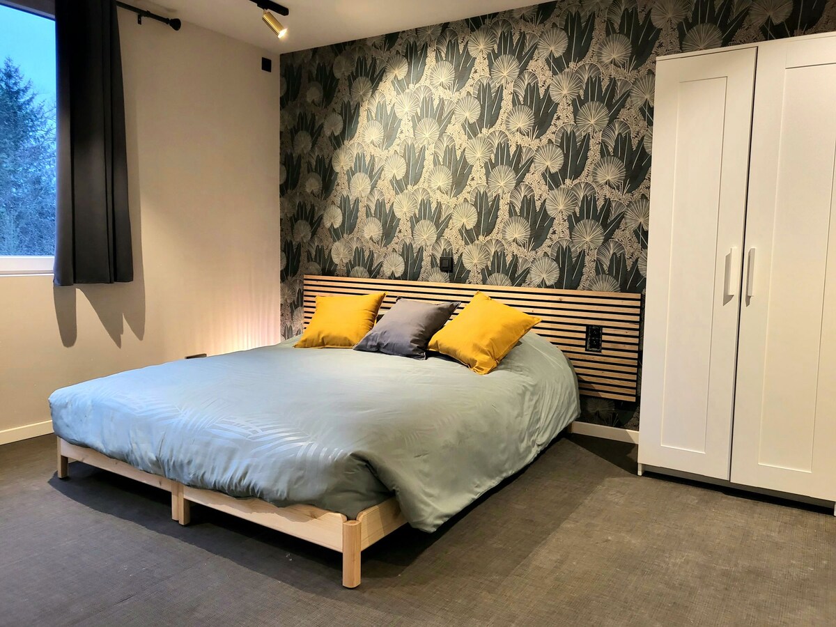 A spacious sleeping area features a large bed with a light blue duvet and two yellow pillows. The wall behind the bed is adorned with a floral wallpaper design, while a two-door wardrobe stands nearby. Natural light enters through a window dressed in dark curtains.