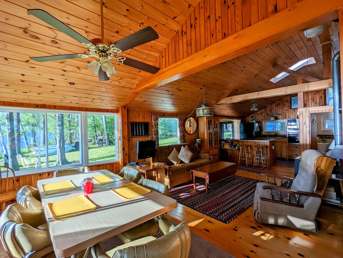 An airy living and dining area features wood paneling and large windows that frame views of the outdoors. A dining table is set with placemats and a red candle, while a comfortable seating area includes a sofa and armchair. Light streams in from overhead skylights.