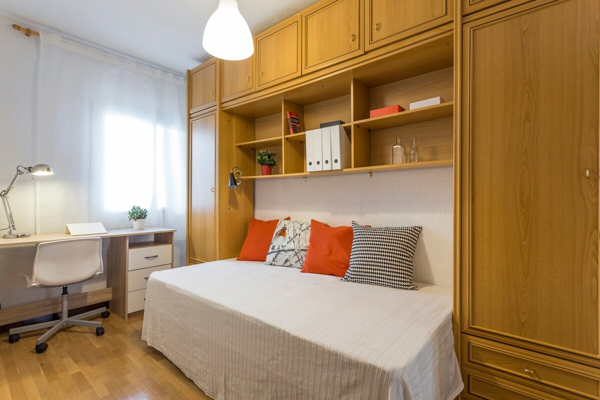 A well-lit room featuring a spacious bed adorned with decorative pillows. A wooden desk with a lamp and a chair is positioned nearby. A wall-mounted shelf holds organized items. Natural light enters through a window covered with sheer curtains, complementing the warm wooden cabinetry.