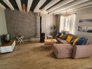 A welcoming living room features a comfortable sectional sofa adorned with colorful cushions. A television is placed on a white media console, and a small wooden table sits nearby. Natural light filters through the windows, illuminating the wooden floors and accentuating the room's cozy decor.