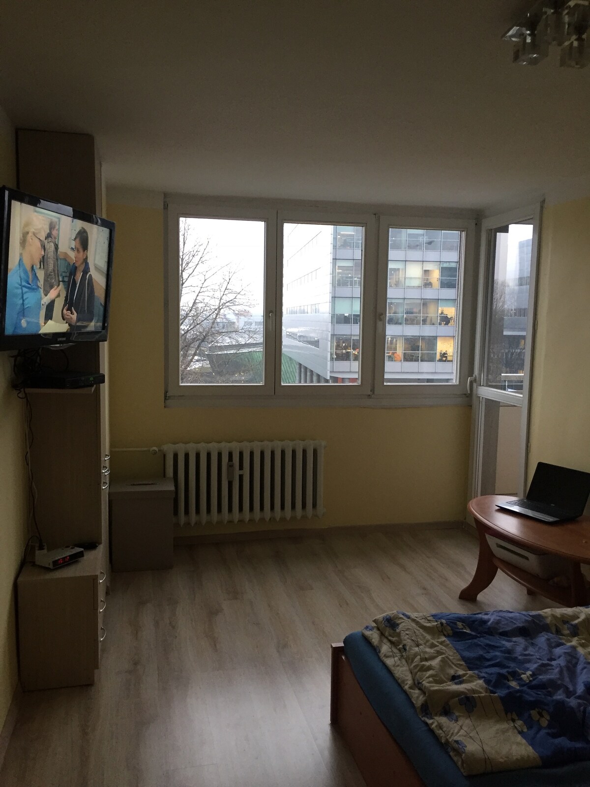 A room features large windows allowing ample natural light, with a view of the surrounding buildings. A wooden table alongside a laptop is present, while a compact television is mounted on the wall. A spacious bed is adorned with a patterned blanket, and a radiator is positioned beneath the windows.
