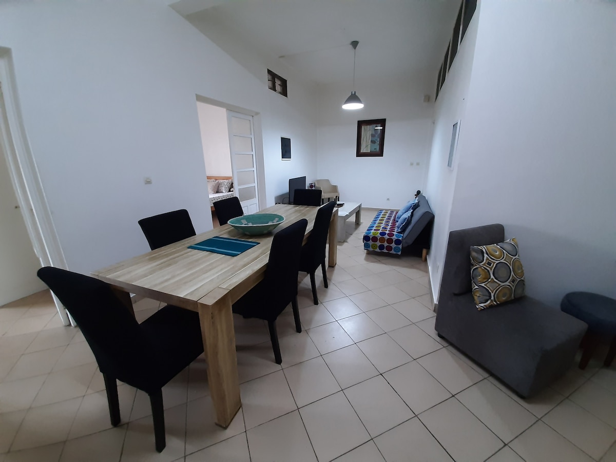 A dining area is presented with a large wooden table surrounded by dark chairs. A small sofa with colorful cushions is visible in the corner, along with a light-colored carpet on the tiled floor. Natural light enters through the open layout, creating a spacious atmosphere.