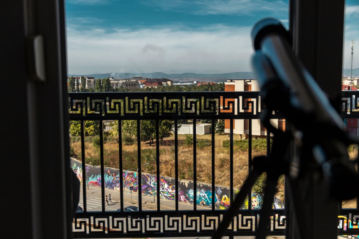 A view from the balcony showcases a vibrant street adorned with colorful art graffiti. A telescope is positioned near the balcony railing, framed by the intricate pattern of the railing. The landscape in the background features distant hills under a clear sky.