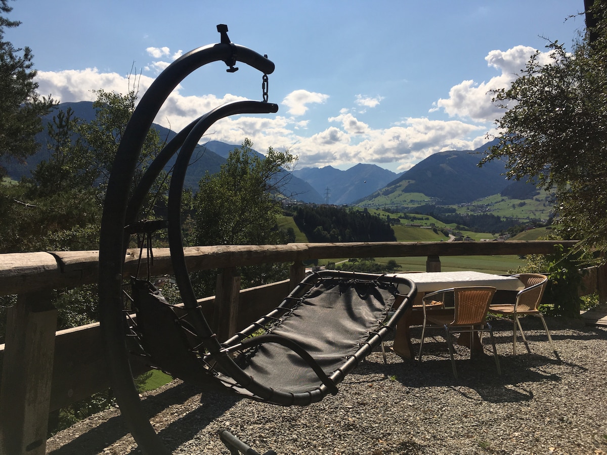 A hanging lounge chair is positioned on a rustic terrace, surrounded by mountains and greenery. A dining table with several chairs is visible nearby, inviting relaxation and outdoor dining. The cloudy blue sky enhances the serene landscape.
