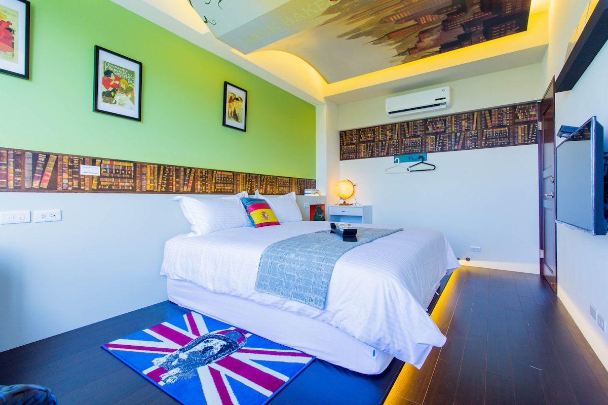 A well-lit guest room features a spacious bed dressed in white linens with an accent blanket. A colorful rug lies underfoot, and framed artwork adorns the walls. An air conditioning unit is mounted, and a wall-mounted television is positioned opposite the bed.