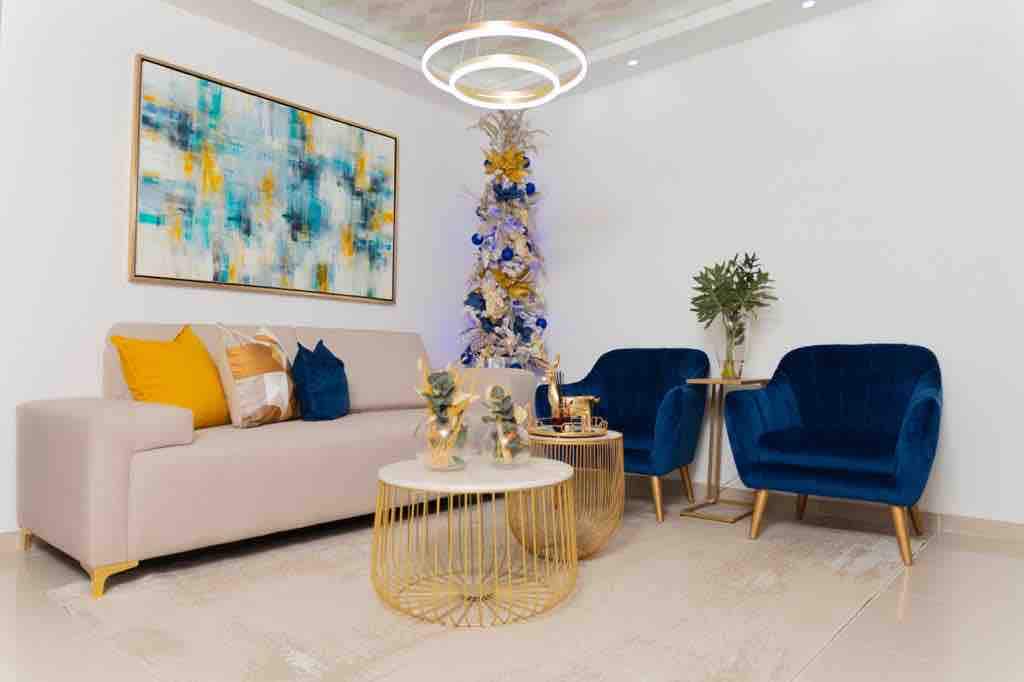 A modern living area features a light-colored sofa adorned with colorful throw pillows, complemented by two plush blue chairs. A round coffee table with a gold base sits at the center, surrounded by decorative items. A stylish Christmas tree adds a festive touch to the space.