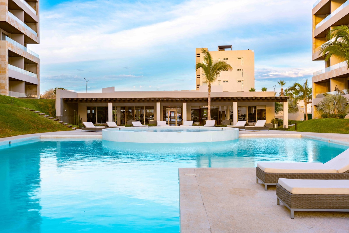 A large pool is centered within the property, surrounded by well-maintained landscaping. Lounging chairs are positioned along the water's edge, and a modern club house is visible in the background, featuring large glass windows that open to the outdoor space.