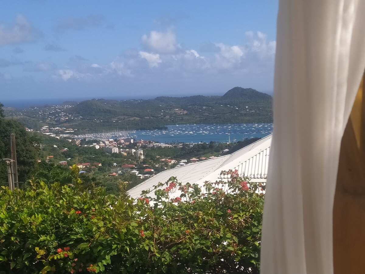 A panoramic view of the scenic coastline is captured, featuring lush greenery in the foreground and a tranquil bay dotted with boats. The sky is bright with clouds, and distant hills create a natural backdrop, showcasing the beauty of the surroundings.