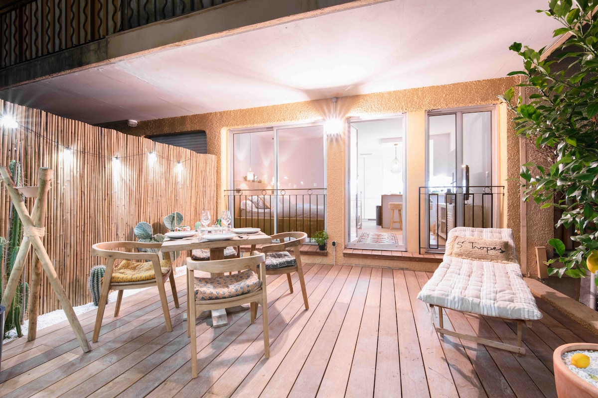 A bright wooden terrace features a dining table set for four and a lounge chair, surrounded by decorative plants and a wooden privacy screen. Large glass doors provide access to the indoor living space, enhancing the connection between inside and outside.