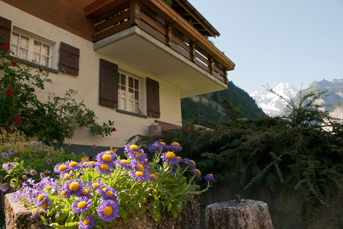 The exterior of the chalet features a balcony with wooden railings, nestled among lush greenery. Vibrant purple flowers bloom in the foreground, complementing the natural landscape, while snow-capped mountains rise in the background under a clear blue sky.