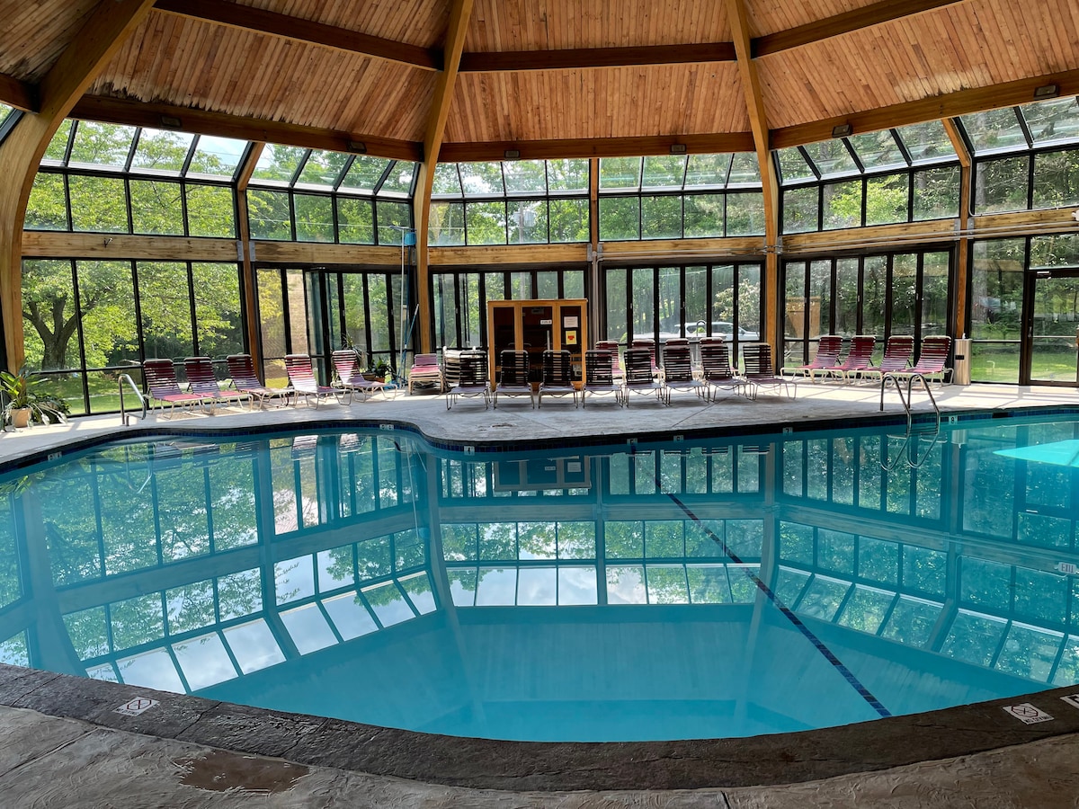 An indoor swimming pool is surrounded by large glass windows, allowing natural light to fill the area. Chaise lounges line the poolside, providing a space for relaxation. The wood-paneled ceiling adds warmth, and reflections can be seen on the water's surface.