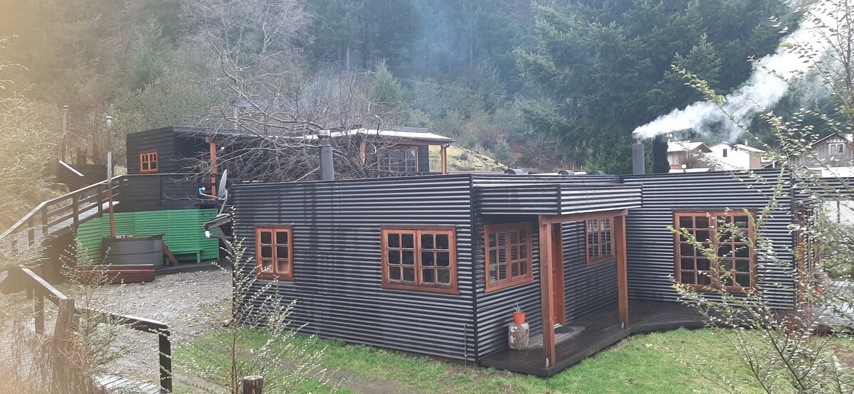 A collection of rustic buildings is nestled within a natural setting, featuring dark wooden siding and warm-toned windows. Smoke gently rises from a chimney, indicating a cozy indoor ambiance. Surrounding greenery adds to the tranquil atmosphere of the property.