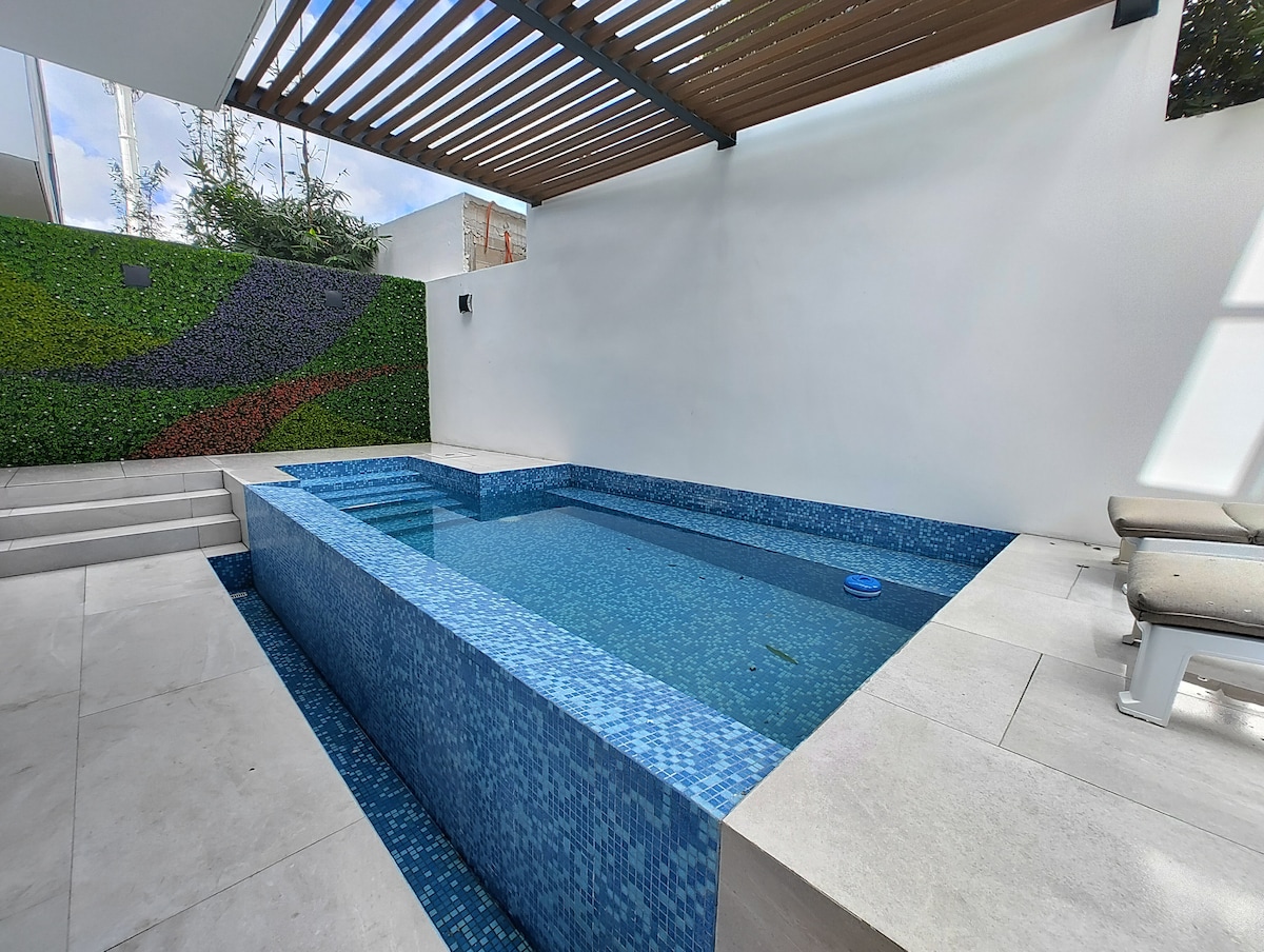 A serene swimming pool features a mosaic of blue tiles, reflecting sunlight. The space is framed by a wooden pergola and a vertical garden wall, adding a touch of greenery. Light-colored tiles surround the pool, creating an inviting area for relaxation.