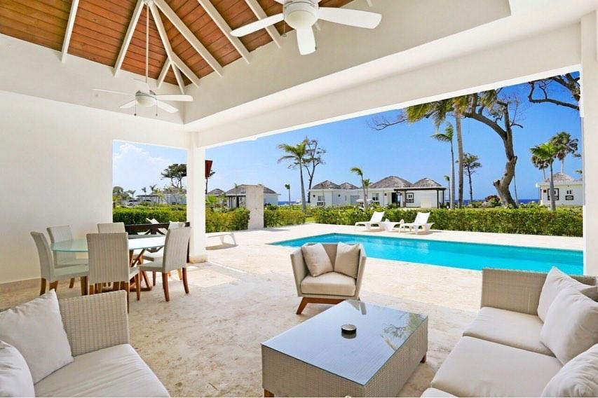An outdoor patio area features a comfortable seating arrangement with a coffee table, surrounded by a dining table for six. The private pool is visible in the background, bordered by lounge chairs and lush greenery, under a clear blue sky.