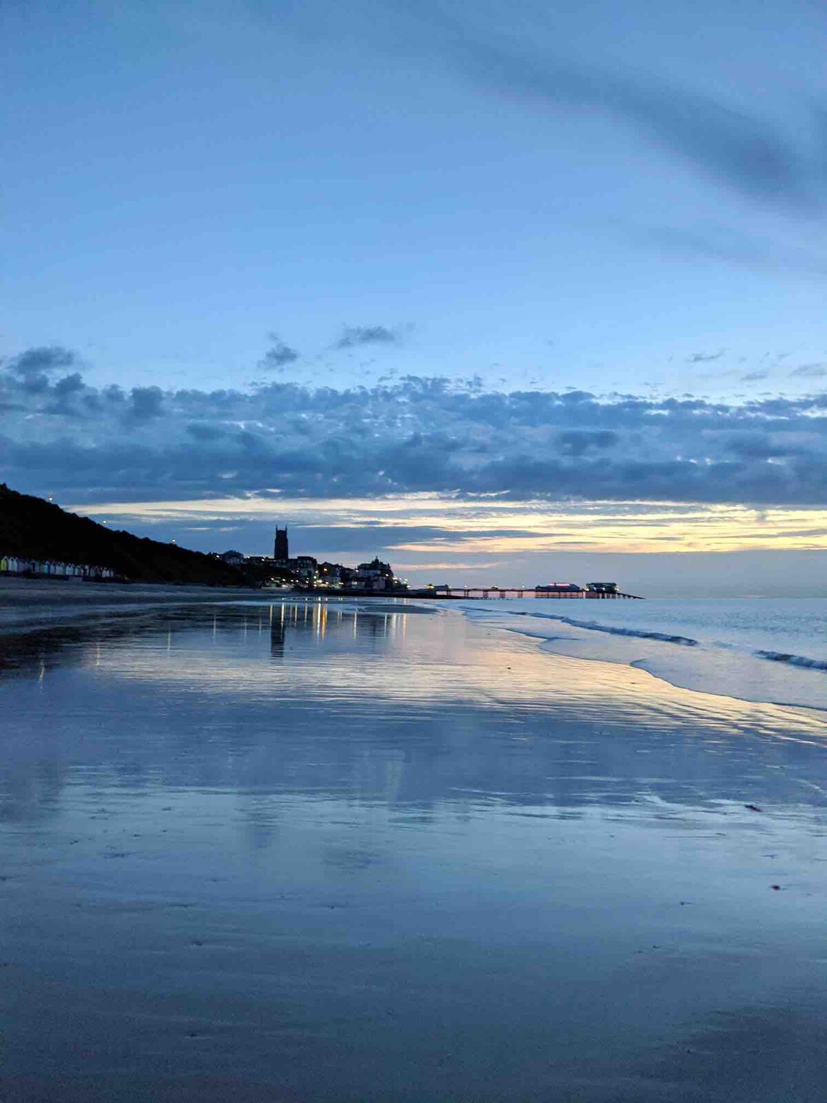A serene beach scene captures the gentle waves reflecting the soft hues of the evening sky. The shoreline glistens under fading light, with distant buildings and a pier visible along the coast, framed by a backdrop of soft clouds.