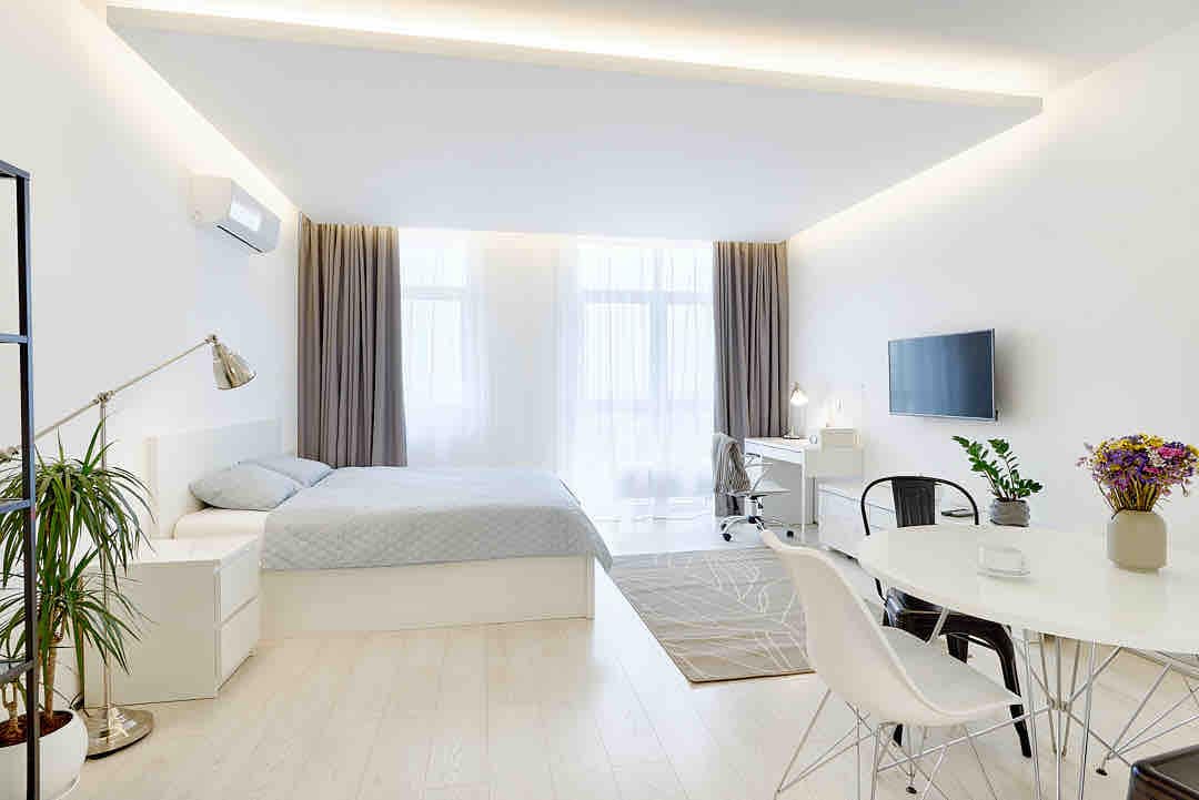 A minimalist studio apartment is displayed, featuring a spacious bed dressed in soft bedding. A work desk with a chair is positioned near a large window, providing natural light. A round dining table is set with stylish chairs, and greenery adds a touch of nature.