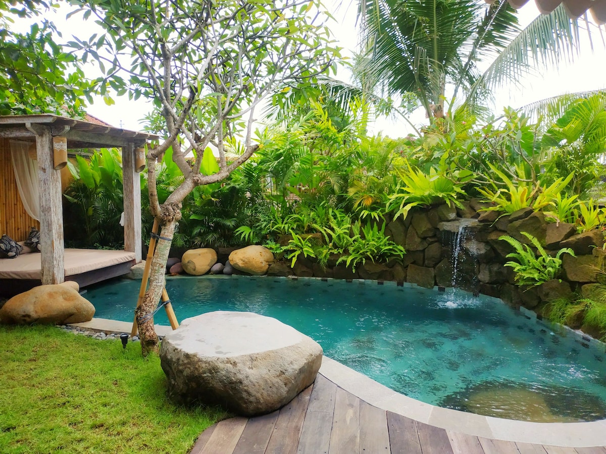 A serene swimming pool is framed by lush greenery, including tall palms and ornamental plants. A natural stone waterfall gently cascades into the pool. A wooden deck and a gazebo can be seen in the background, providing shaded areas for relaxation.