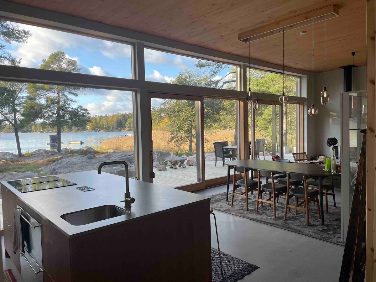 An open-plan kitchen and dining area features large windows that provide views of the surrounding nature and water. A modern kitchen island is complemented by a wooden dining table set for six. Minimalist pendant lights hang from the ceiling, enhancing the spacious atmosphere.