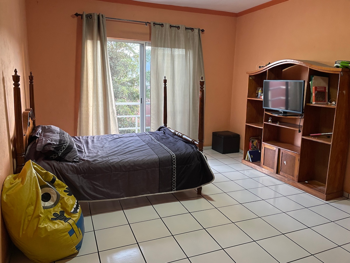 A bright room features a wooden bed with dark bedding and a colorful bean bag. A wooden entertainment unit holds a television, and natural light enters through large sliding doors covered with sheer curtains, providing a view of greenery outside.