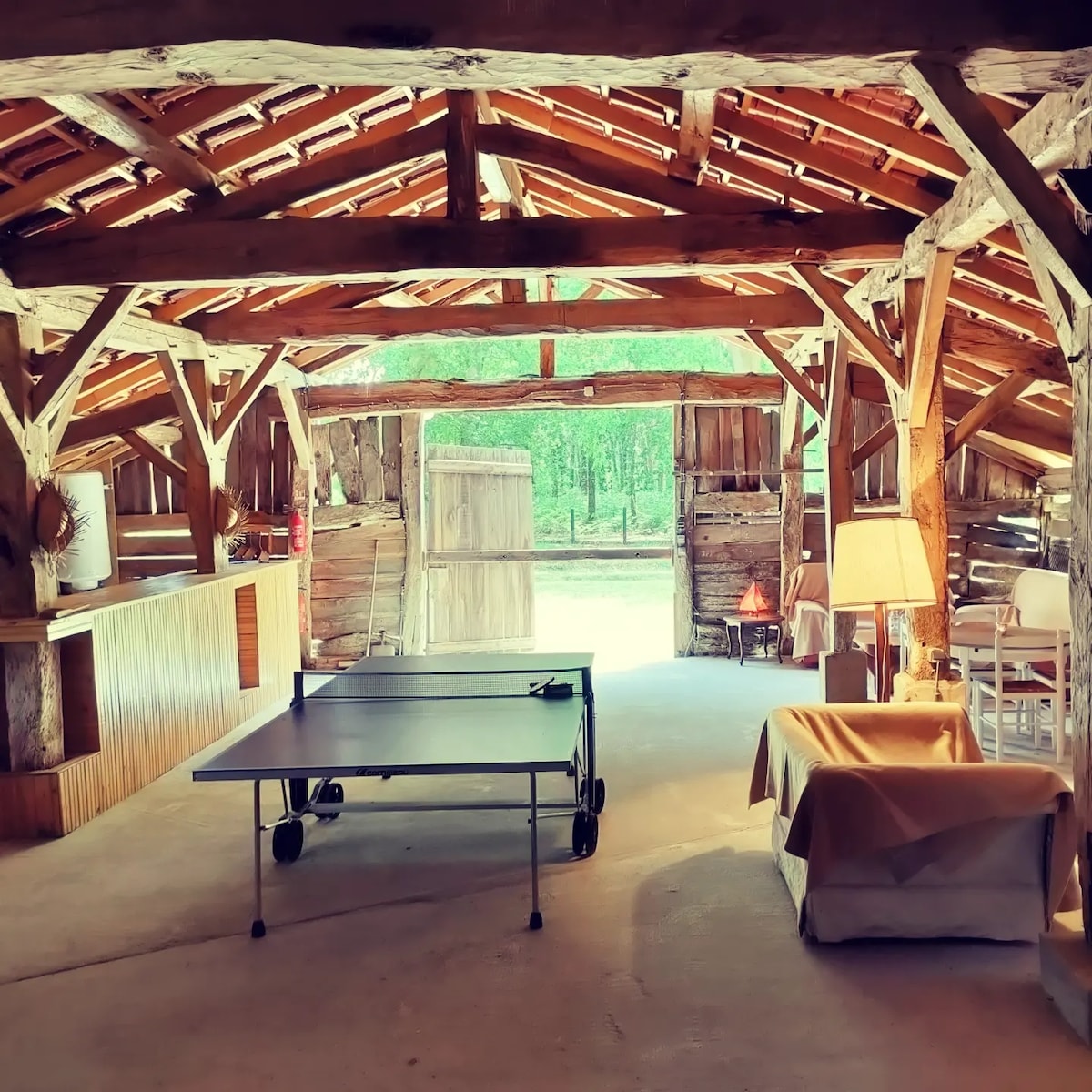 A rustic common space features exposed wooden beams and a welcoming atmosphere. A ping pong table occupies the center, while a comfortable seating area is positioned nearby. Sunlight streams through open doors, illuminating the natural surroundings visible through the entrance.