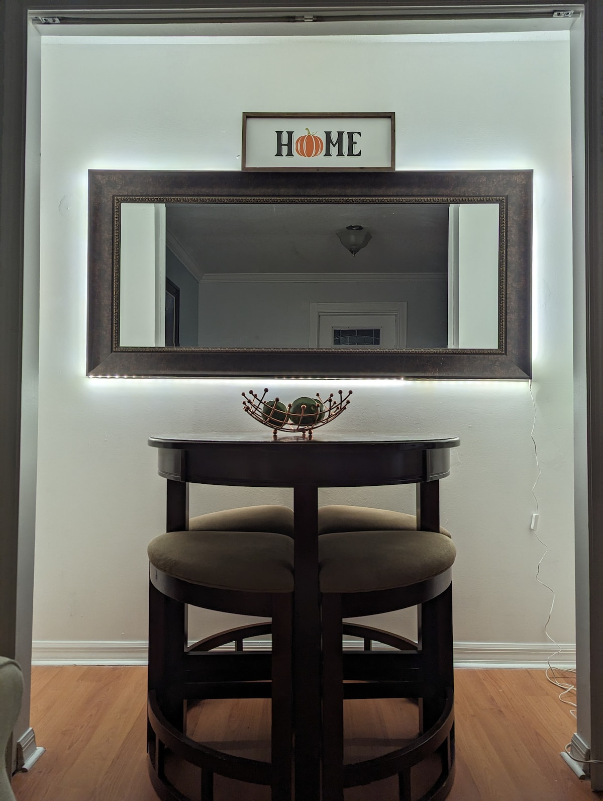 A cozy seating area is highlighted by a circular table and two cushioned stools. A large, illuminated mirror reflects light, enhancing the space. Above the mirror, a decorative sign with the word 'HOME' adds a personal touch.