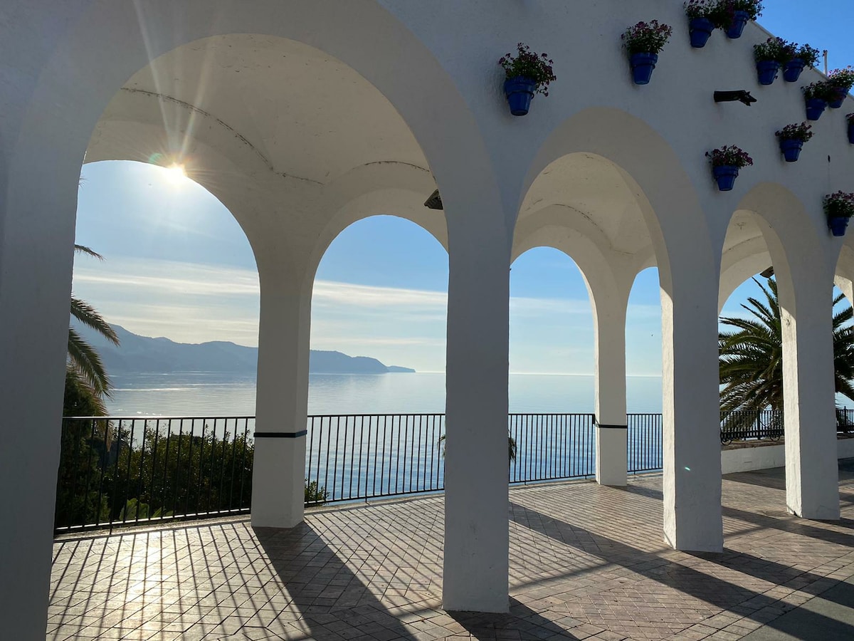 An open terrace is framed by three large arches, revealing expansive sea views. Potted flowers hang along the top of the arches, adding a touch of color. A tiled floor reflects the sunlight, enhancing the tranquil atmosphere of the elevated location.