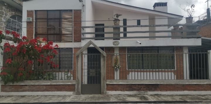 A two-story building is displayed, featuring a combination of brick and white exterior walls. A metal gate provides entrance, and vibrant red flowers enhance the outdoor space. The upper level includes a balcony with a railing, while large windows allow ample natural light.