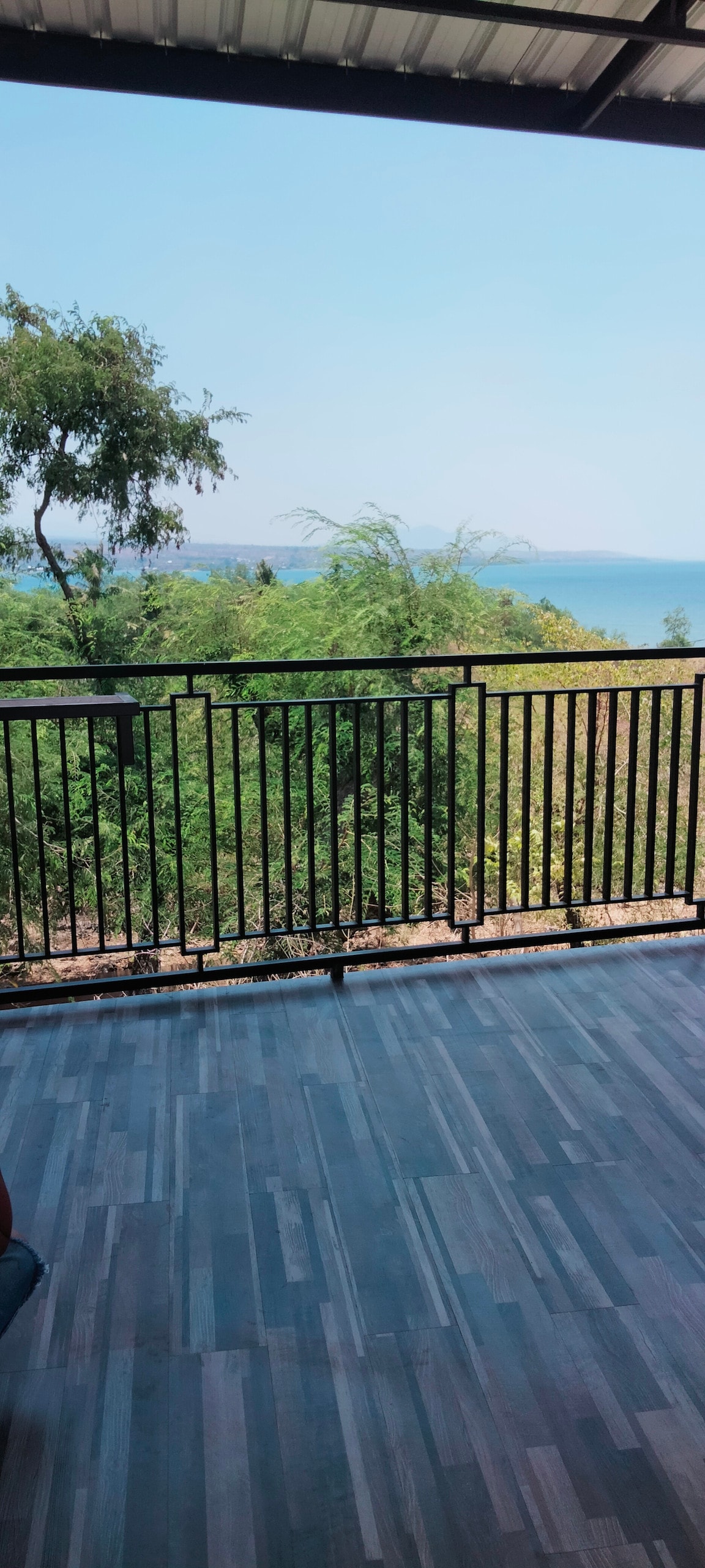 A balcony area is shown with a metal railing offering a view of lush greenery and a distant body of water. The flooring blends wood-like tiles, creating a modern and clean appearance, while natural light is evident in the open space.