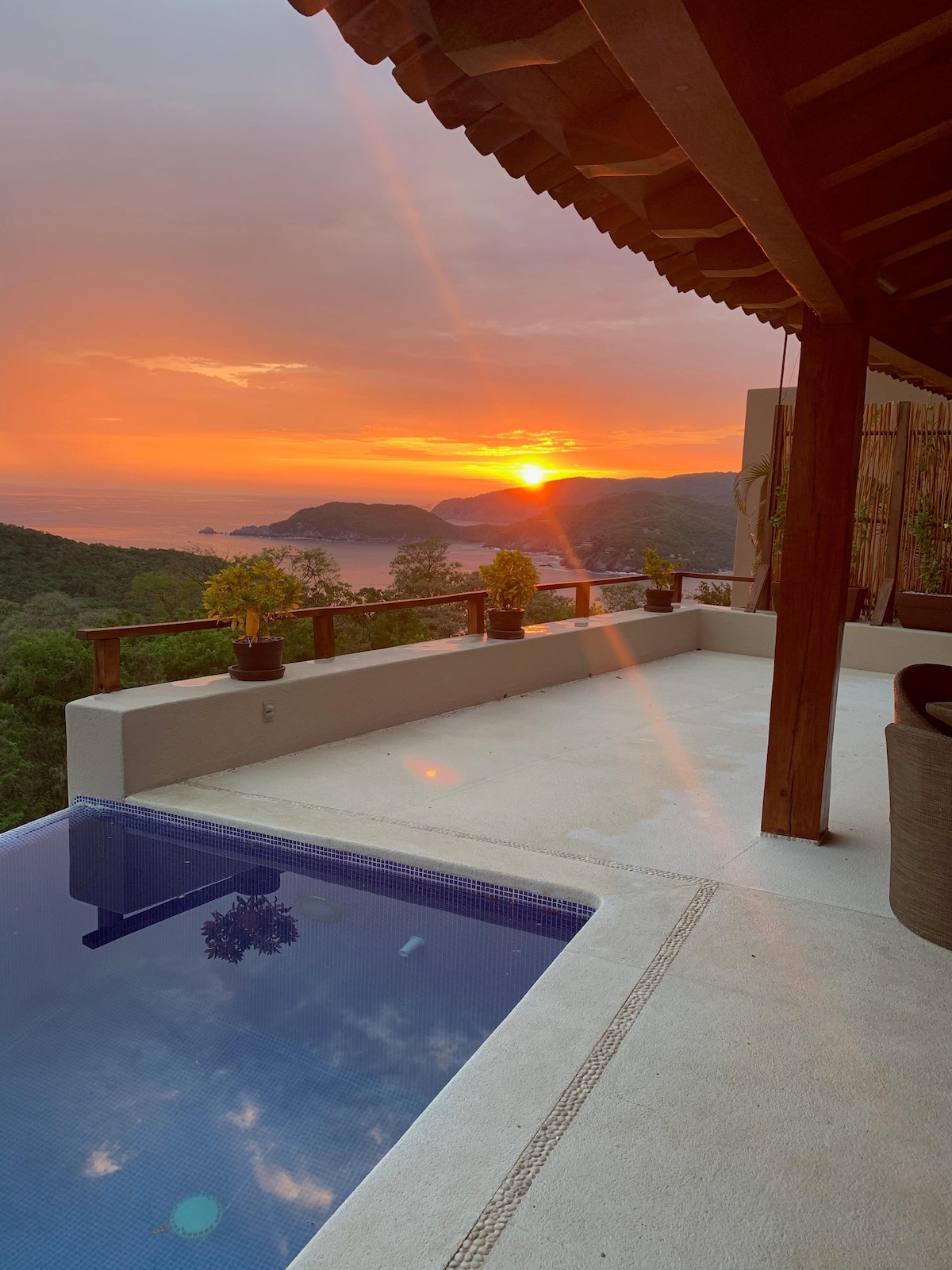 An outdoor terrace features a private infinity pool, framed by wooden posts and potted plants. A breathtaking sunset over the ocean is visible in the distance, casting warm hues across the sky and mountains.