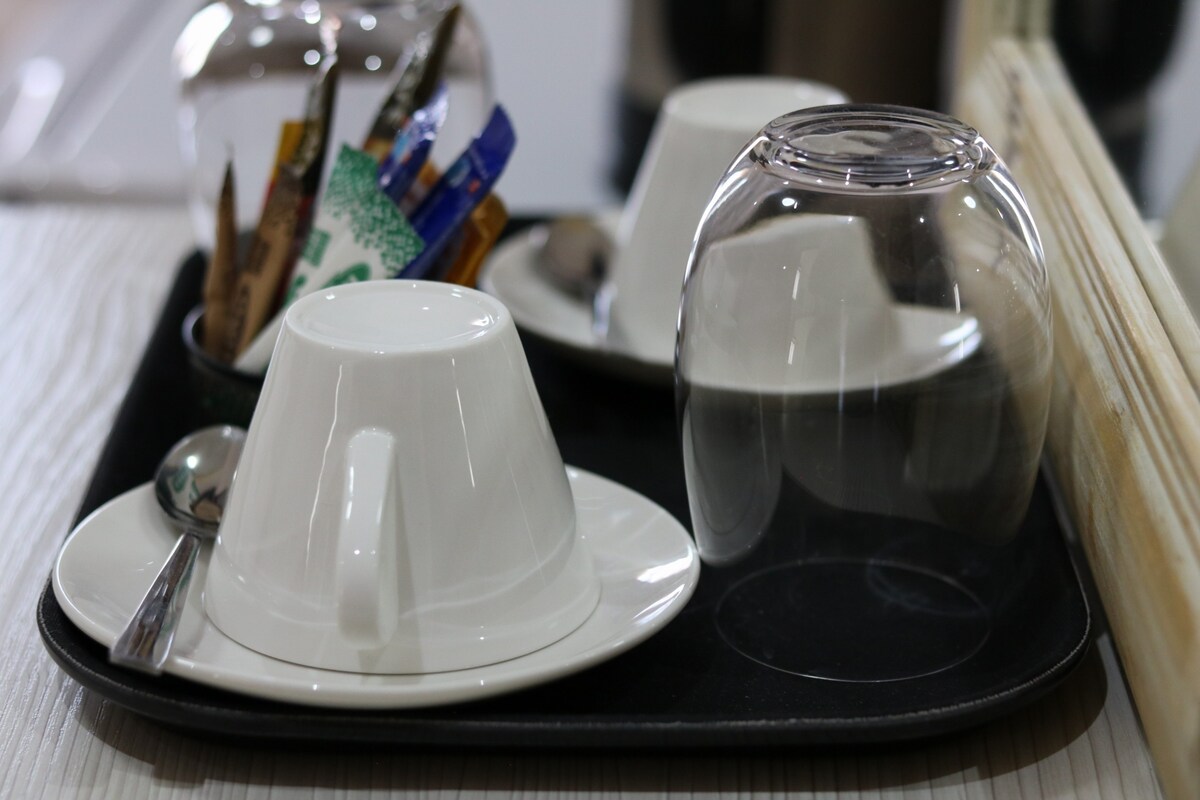A tray is set with two white cups on matching saucers, accompanied by a metal spoon. An assortment of sugar packets and stirrers is organized in a glass container, which is protected by a transparent cloche. The soft lighting creates a warm ambiance.