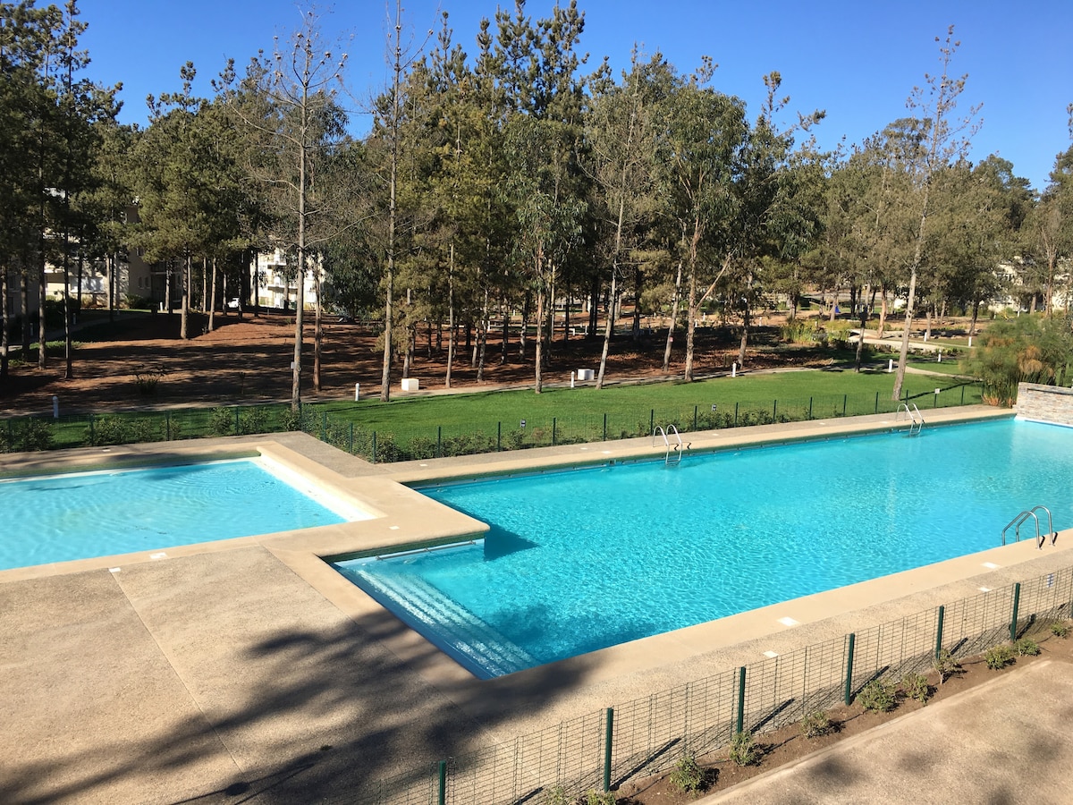 A spacious outdoor pool area is surrounded by greenery, with two swimming pools positioned side by side. One pool is designed for children, while the other serves as a lap pool. Trees and landscaped areas are visible in the background, creating a tranquil environment.