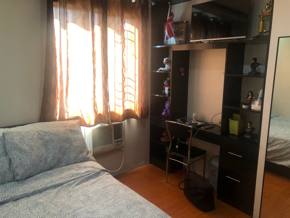 A cozy bedroom is shown with a spacious bed covered in a patterned duvet. Natural light filters through partially closed curtains, illuminating a small desk with a chair and various decorative items. An air conditioning unit is positioned under the window, enhancing comfort.