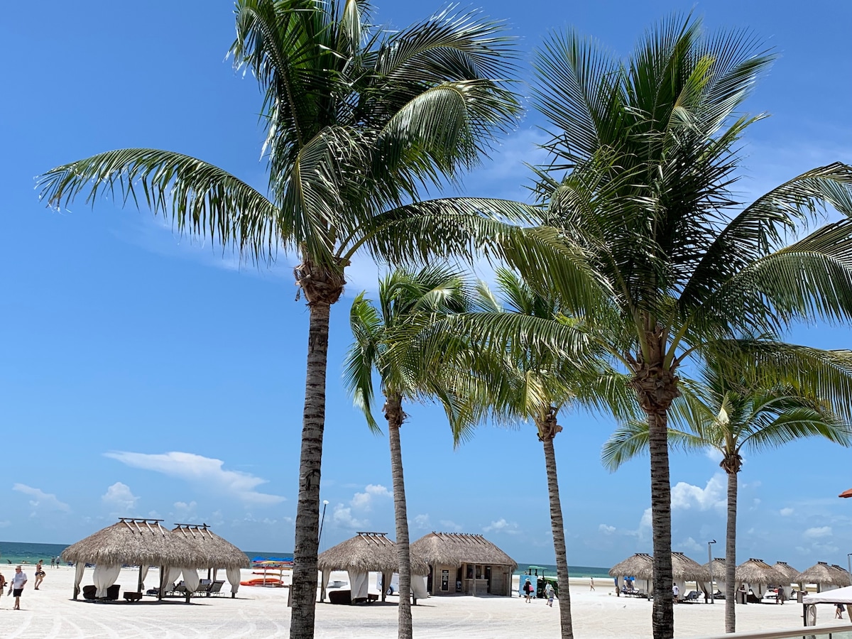 Palm trees sway gently above a sandy beach, where thatched-roof cabanas provide shade. A clear blue sky stretches overhead, complemented by a distant horizon meeting the tranquil sea. Beachgoers can be seen strolling along the waterfront, enjoying the warm, sunny day.