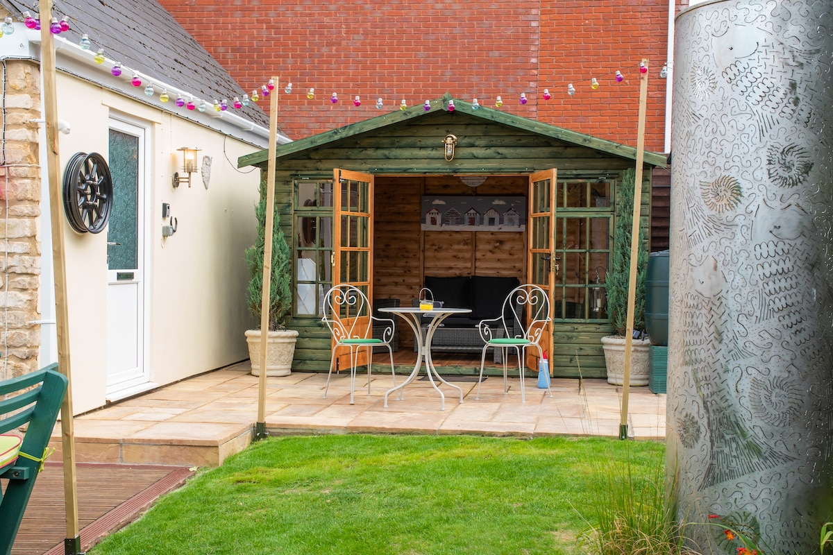 A charming summerhouse is featured with open double doors revealing a small table and two chairs, positioned on a tiled patio. Decorative lights are strung across the top, enhancing the inviting outdoor space surrounded by grass and potted plants.