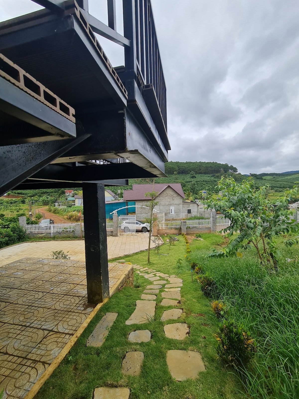 The image features a pathway made of stone leading through lush greenery. A partial view of a house is visible in the background, with a cloudy sky above. The structure above creates an interesting perspective, highlighting the spacious outdoor area below.