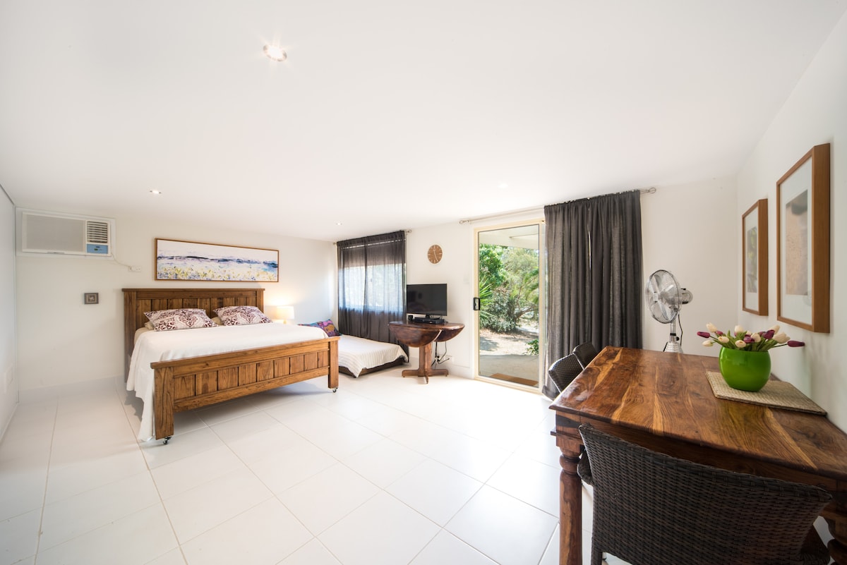 A spacious guest suite is presented with light-colored tiled flooring. A king-sized wooden bed is paired with a single trundle bed in a corner. A wooden table with woven chairs and a TV are visible. Large windows allow natural light to fill the room.