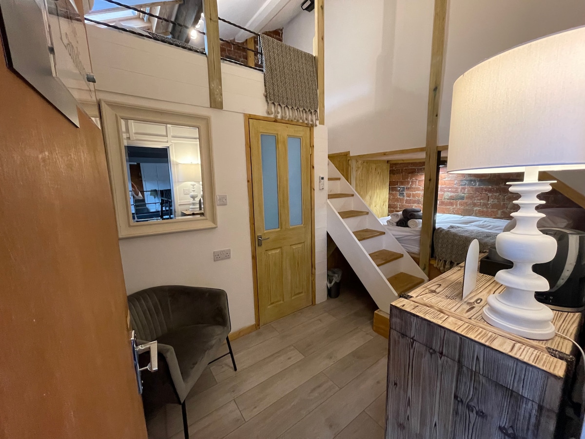 An entrance area features a small chair beside a wooden console table. A decorative lamp provides illumination. The room is highlighted by a mirror and a door leading to the upper sleeping space. Wooden beams and brick accents add character to the warm atmosphere.