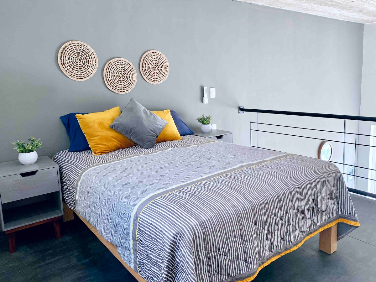 The sleeping area features a spacious bed dressed in a striped blanket with decorative pillows in vibrant shades. Light-colored wall accents complement the gray walls, and wall-mounted decor adds texture. Nightstands with small plants are positioned on either side of the bed.
