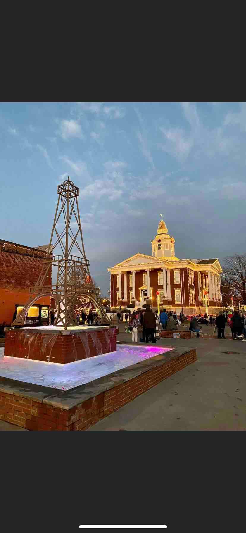 A historic building with a prominent clock tower is featured, highlighted by warm evening lighting. People gather around an illuminated fountain adorned with a decorative metal structure. The sky exhibits soft hues, complementing the festive atmosphere of the scene.