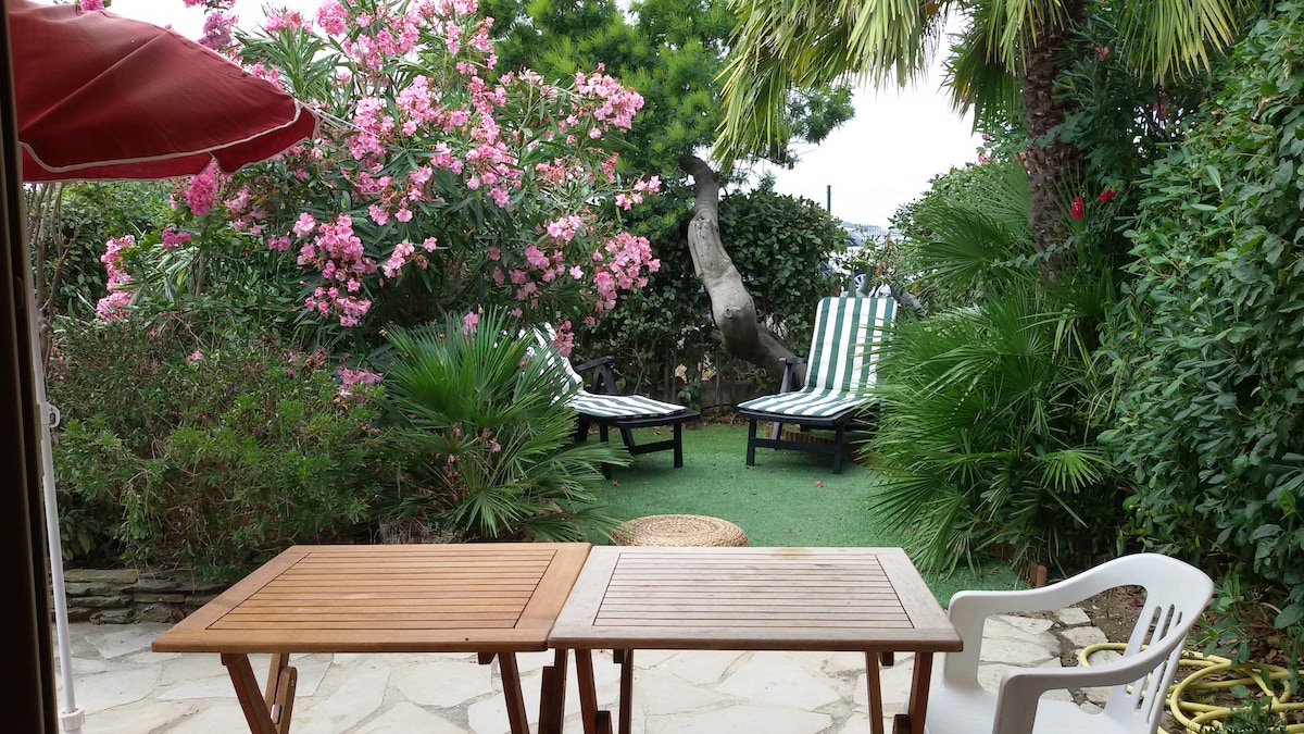 A private garden area is displayed, featuring lush greenery and vibrant pink flowering plants. Two lounge chairs are positioned among the foliage, inviting relaxation. A wooden table is set for outdoor dining, complemented by a white chair. The space is surrounded by tropical plants, providing a serene atmosphere.