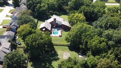 An aerial view showcases a spacious property surrounded by trees, featuring a private swimming pool at the center. The backyard includes a patio area with seating, offering a secluded and tranquil environment. Nearby residential structures are visible, enhancing the sense of privacy.