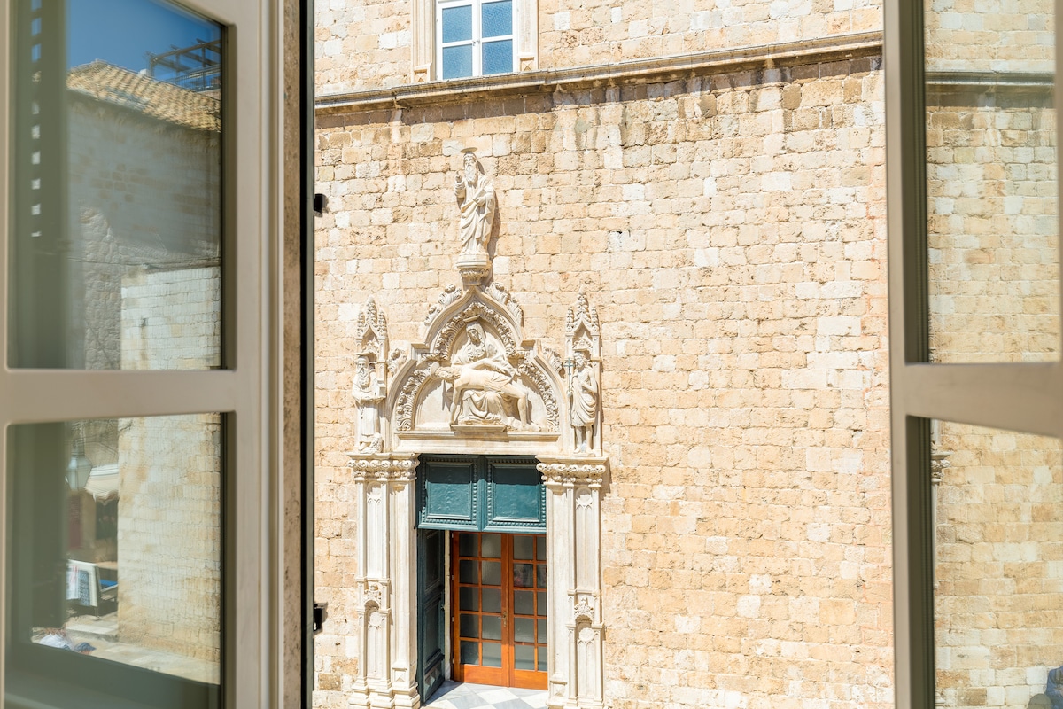 The view from the large windows showcases the historic Franciscan monastery's intricate Gothic Portal, framed by the light stone walls of the building. The detail of the architectural design is clearly visible, highlighting the craftsmanship of the era.