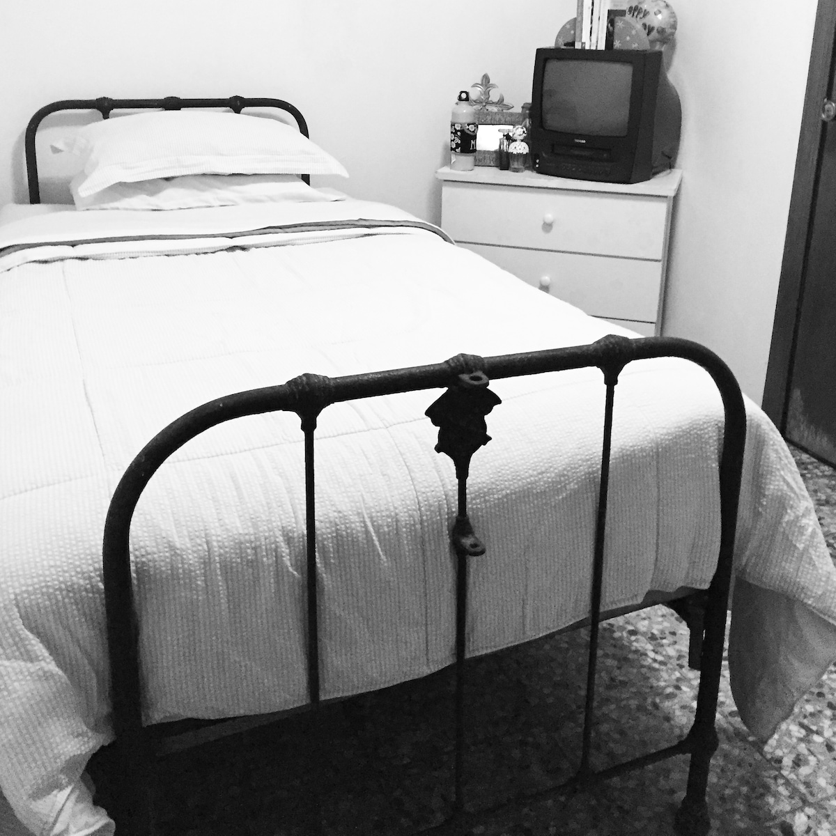 A spacious bed with a white bedspread is positioned in a cozy room. A vintage-style television sits atop a dresser next to a small table adorned with decorative items. Light colors and contrasting textures create a calm environment.