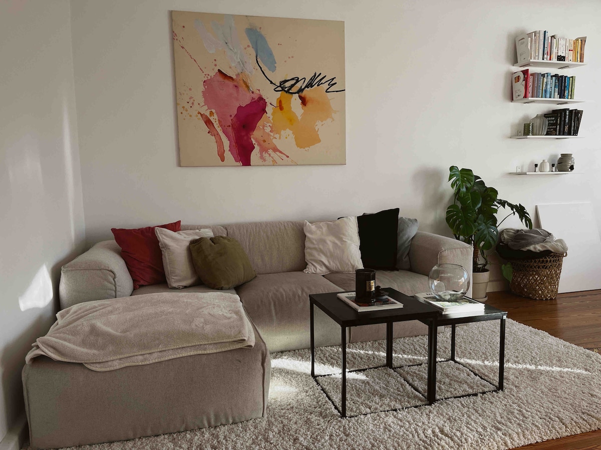 A cozy living area features a light-colored sectional sofa adorned with various cushions. A colorful abstract painting hangs on the wall, while a small coffee table is placed in front. Natural light illuminates the space, highlighting a bookshelf and a potted plant in the corner.