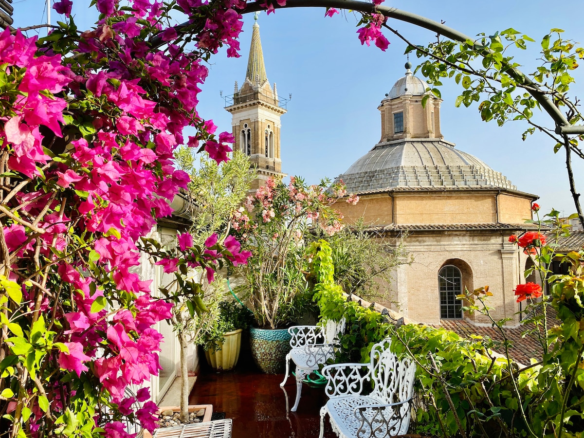 A serene view from the terrace features vibrant pink bougainvillea cascading over a decorative railing. In the background, a domed building and tall steeple are visible against a clear blue sky, enhancing the atmosphere of tranquility.