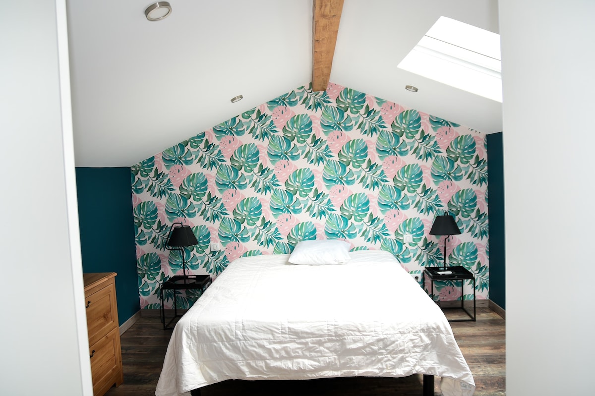 A bright sleeping area features a spacious bed against a vibrant wall adorned with tropical leaf patterns. Windows above allow natural light to illuminate the room, which includes two bedside tables with lamps and a wooden dresser, enhancing the overall design.