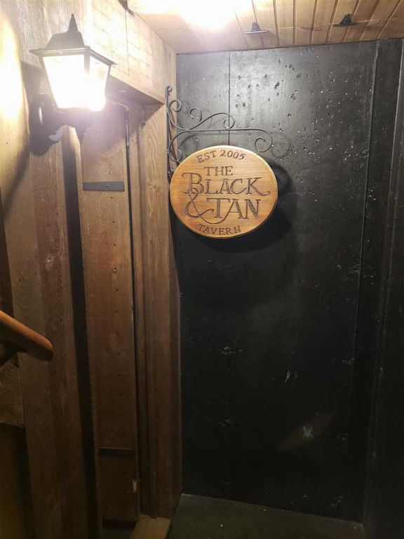 An entrance is depicted with a wooden sign hanging on a black wall, indicating 'The Black & Tan Tavern' established in 2005. A warm light fixture illuminates the area, enhancing the rustic wooden details of the staircase leading to the entrance.