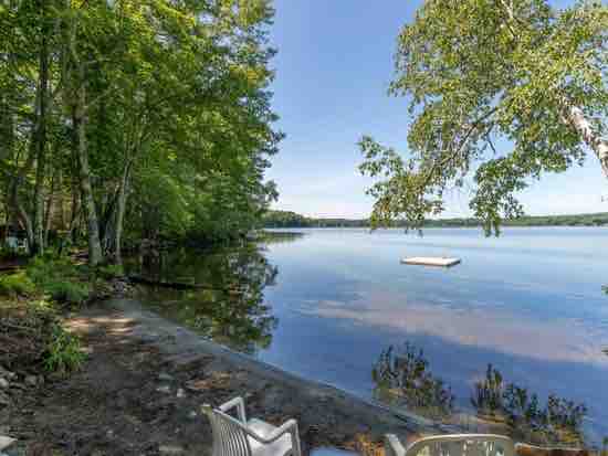 A serene lakeside view is captured, showcasing calm waters reflecting the surrounding greenery. A small dock is visible in the distance, while a pair of chairs sit invitingly on the shore, located under the shade of leafy trees.